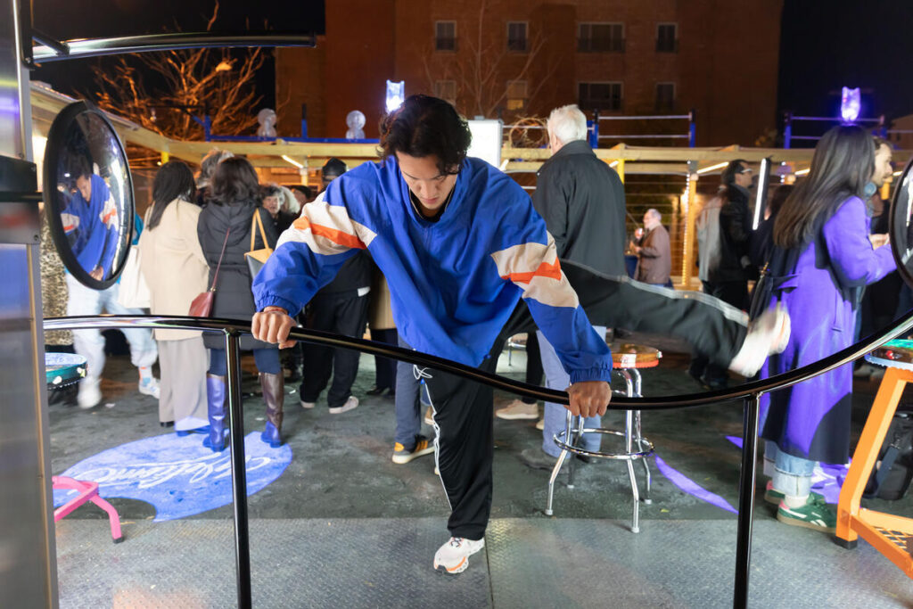 A person in a blue jacket balances on a curved bar, stretching one leg, at an outdoor event at night, with people and lights in the background.