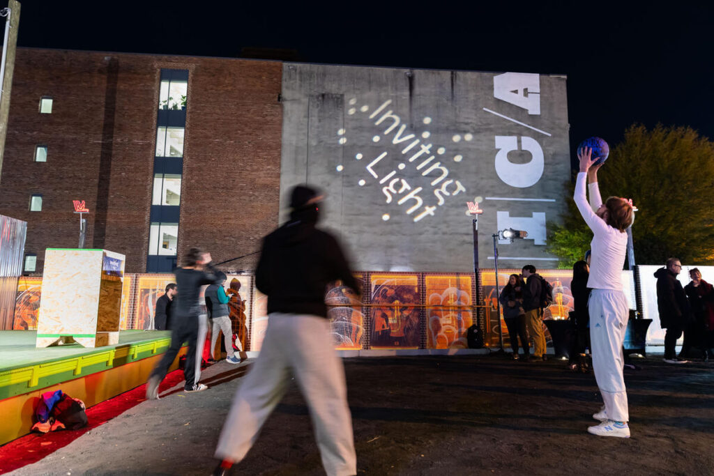 People play basketball outside at night near a building with “Inviting Light” projected on the wall. Bystanders watch, and string lights illuminate the area, creating a festive atmosphere.