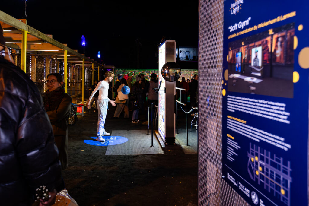 A person stands on a lit circle outdoors at night, with people watching nearby. There are colorful lights, a mural in the background, and an informational sign in the foreground about an exhibit called 6th Gym.