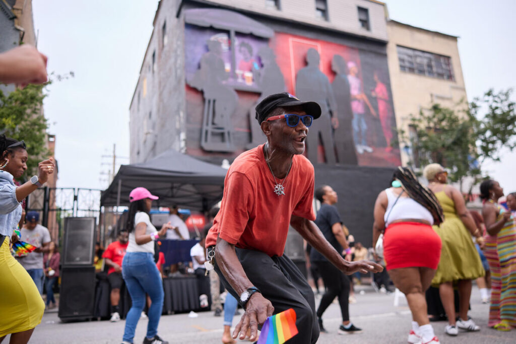 A man in a red shirt and sunglasses dances in the street, holding a small rainbow flag, surrounded by people at an outdoor event with a mural in the background.