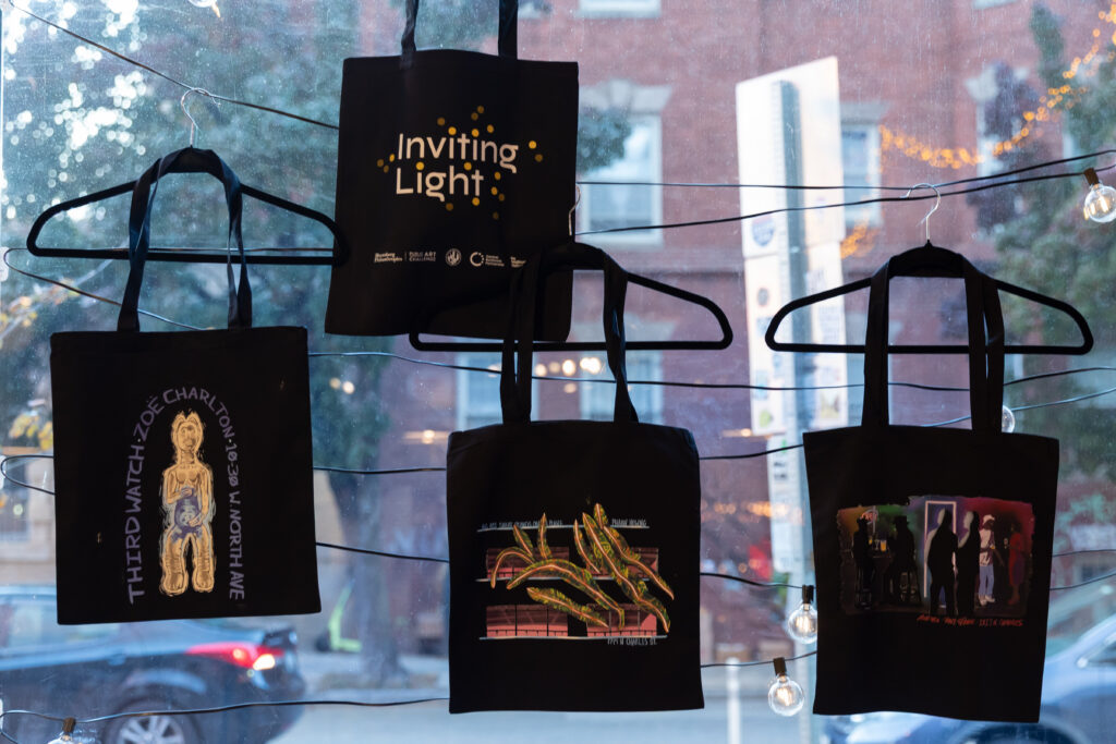 Four black tote bags with different artistic designs and text hang on black hangers in front of a window, with blurred outdoor scenery and string lights visible in the background.