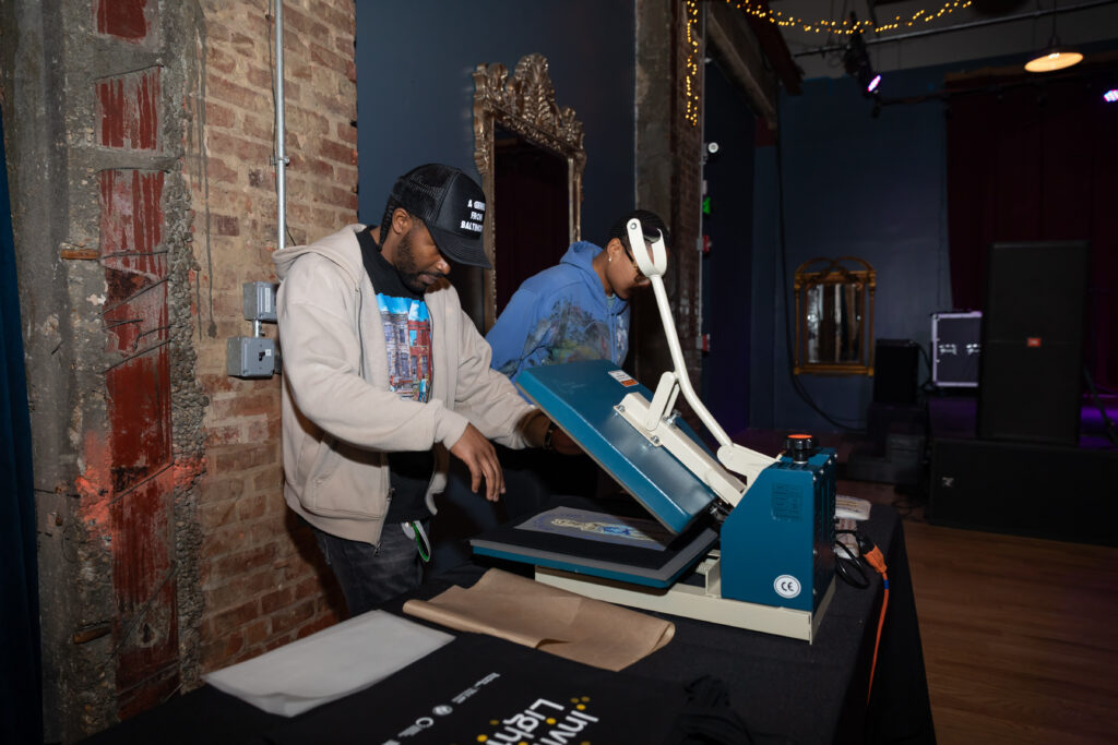 Two people use a heat press machine on a table with t-shirts in a room with exposed brick walls, a mirror, and stage lighting. One person presses the machine while the other stands nearby.