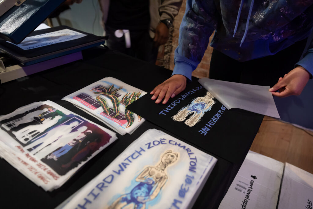 A person arranges printed T-shirts and fabric sheets with various designs and the text THIRD WATCH ZOË CHARLTON on a table, preparing or displaying them in an indoor setting.