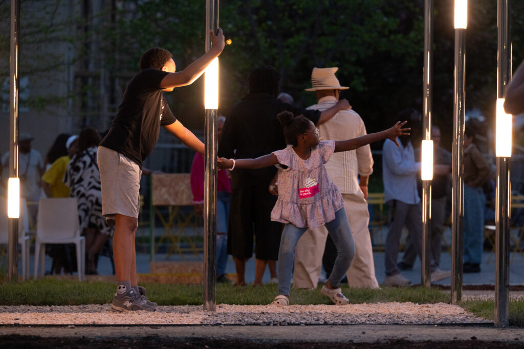 Two children playfully reach for each other around a light pole at dusk, surrounded by people standing and sitting outdoors. Bright lights illuminate the scene.