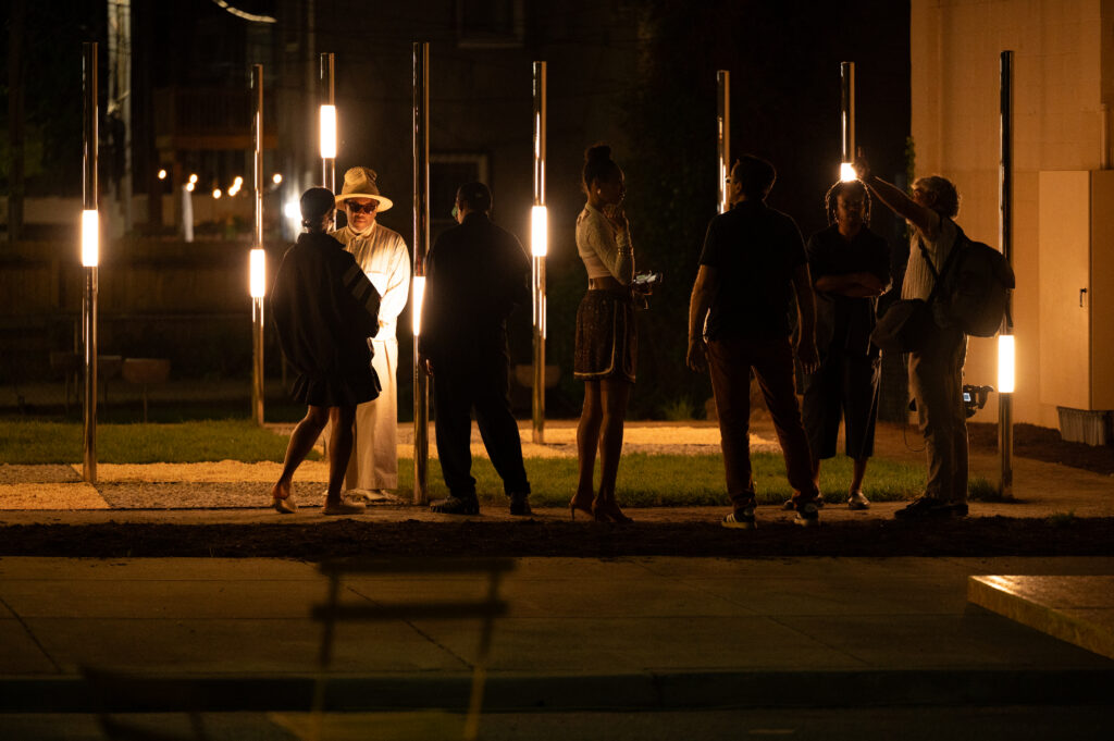 A group of people stands outdoors at night among the vertical light poles of Harmony Park, some talking while others watch, creating illuminated silhouettes on a grassy area near a sidewalk