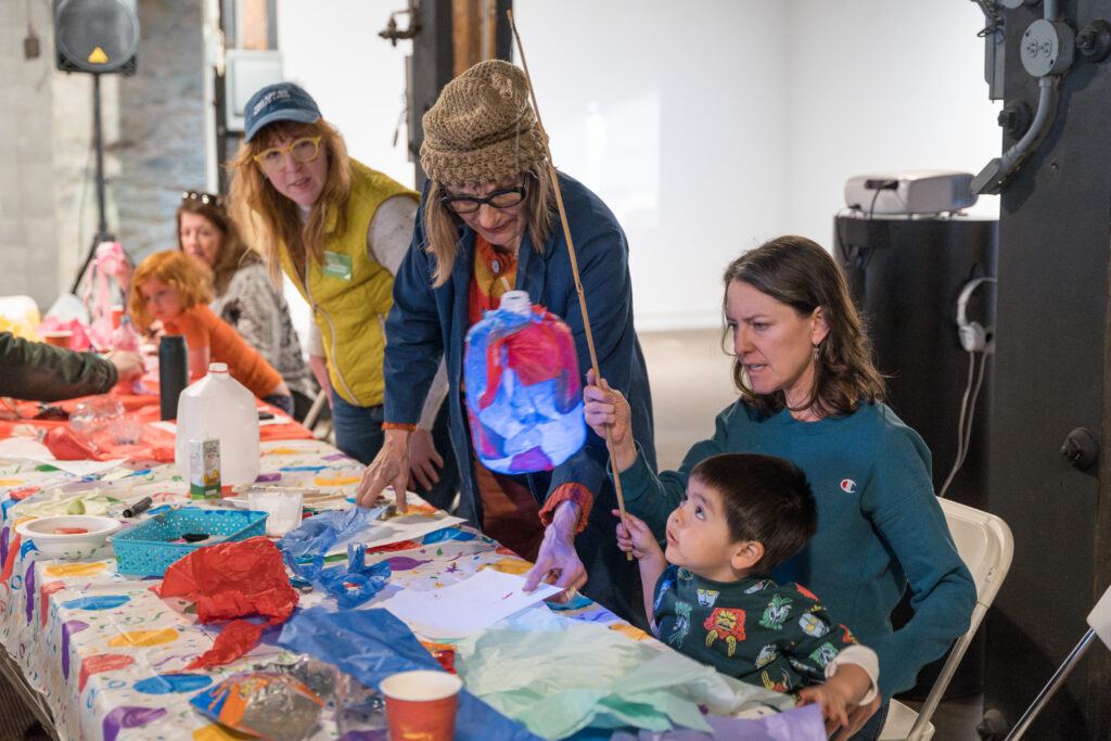 Several adults and a child sit around a colorful table, creating crafts with tissue paper and other materials. An adult helps the child hold up a lantern-like project. The scene is lively and creative.