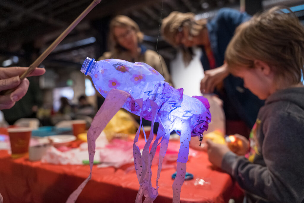 A glowing jellyfish craft made from a plastic bottle and tissue paper is held on a stick. People, including a child, work on crafts at a table covered in red paper in a busy, indoor setting.