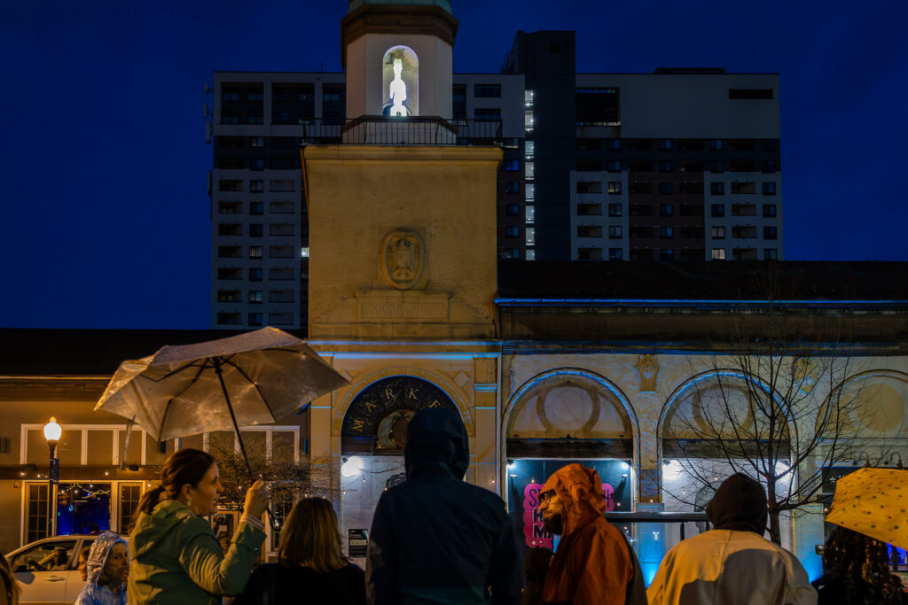 A group of people, some with umbrellas, stand outside at night facing a lit building with a clock tower and statue, with a tall apartment building in the background.