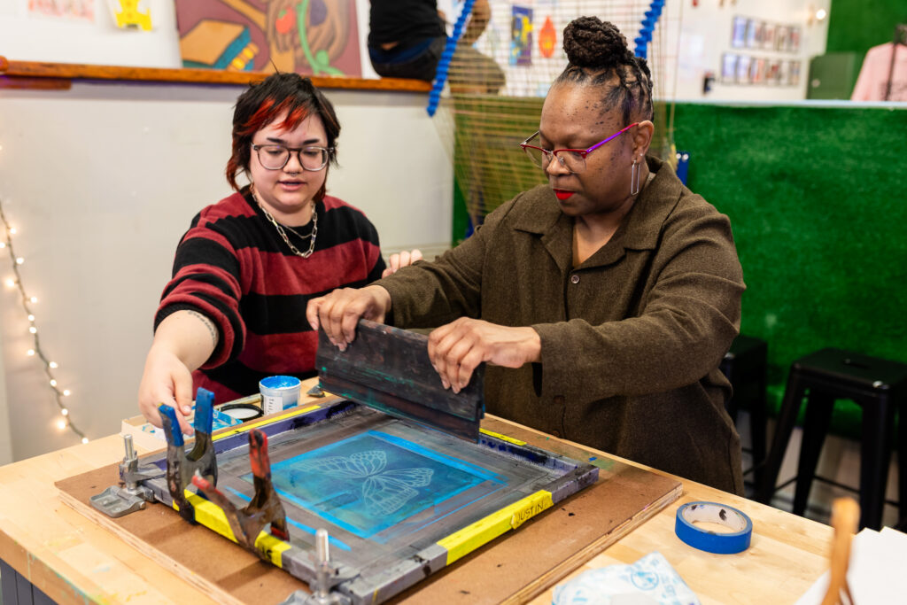 Two people work together on a screen printing project, pressing ink through a stencil onto fabric. One person guides the ink squeegee while the other assists, surrounded by art supplies and colorful decor.