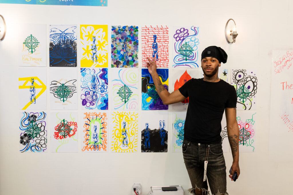 A man in a black shirt and hat points to colorful artwork displayed on a wall, featuring abstract patterns, human figures similar to the pregnant figure in Third Watch, and vibrant designs