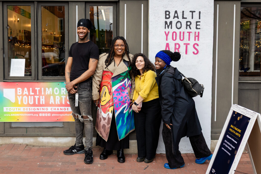 Four people smile while standing outside the Baltimore Youth Arts building, one holding a colorful painted banner. The group stands in front of signs and windows displaying the organizations name.